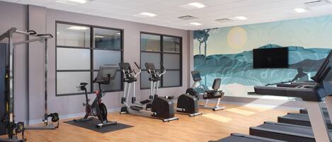 A gym with exercise equipment, large windows, and a mural on the wall.