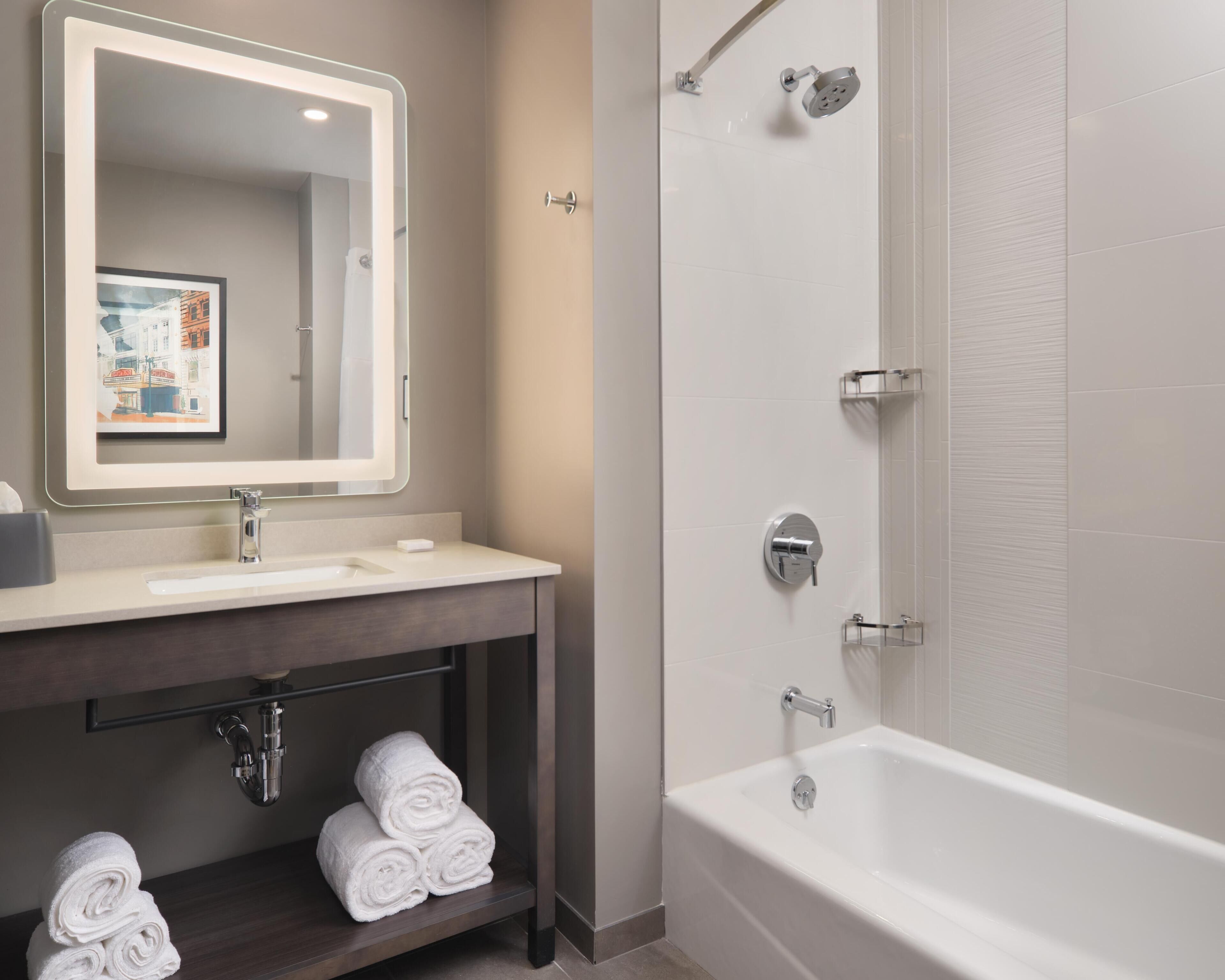 A modern bathroom with a white bathtub, a sink with a mirror, and a shelf with rolled towels.