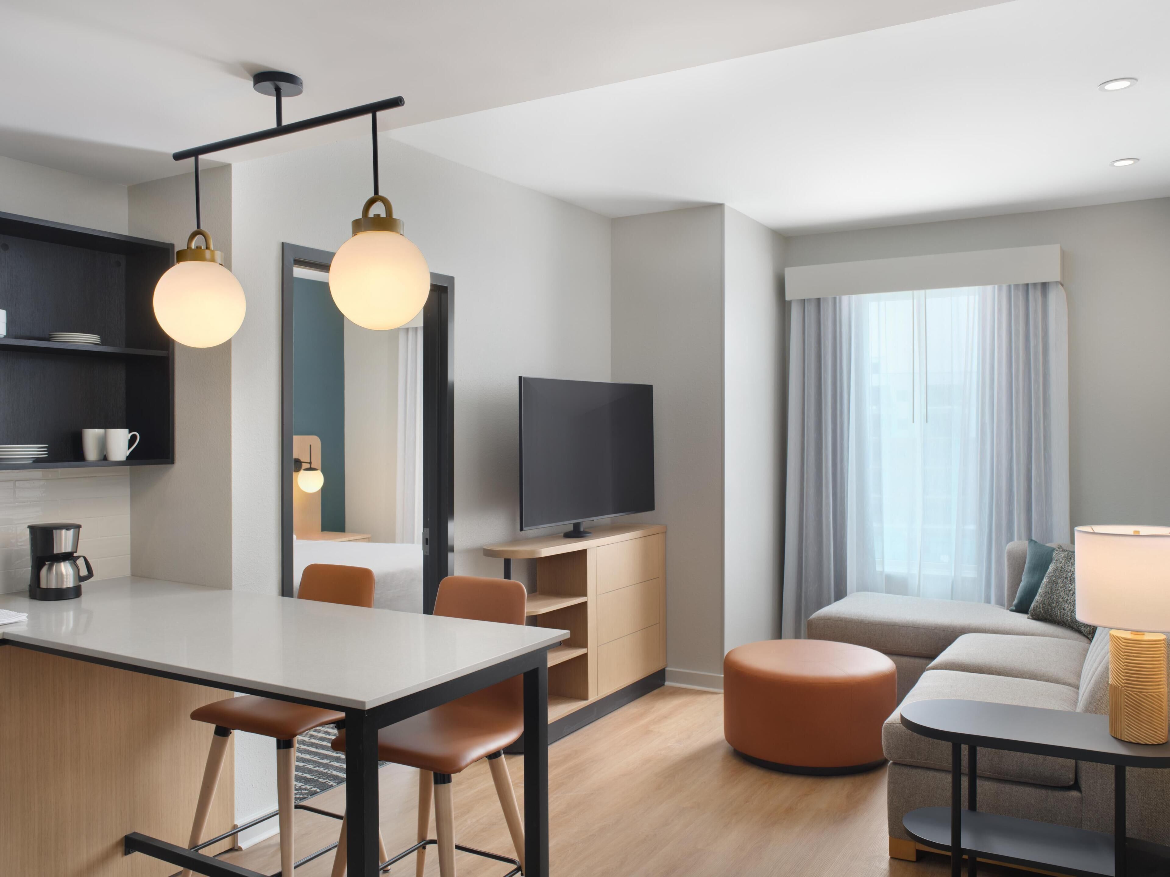 A modern hotel room with a dining area, a flat-screen TV, a sofa, and a lamp.