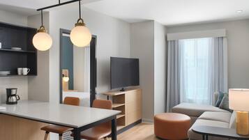 A modern hotel room with a dining area, a flat-screen TV, a sofa, and a lamp.