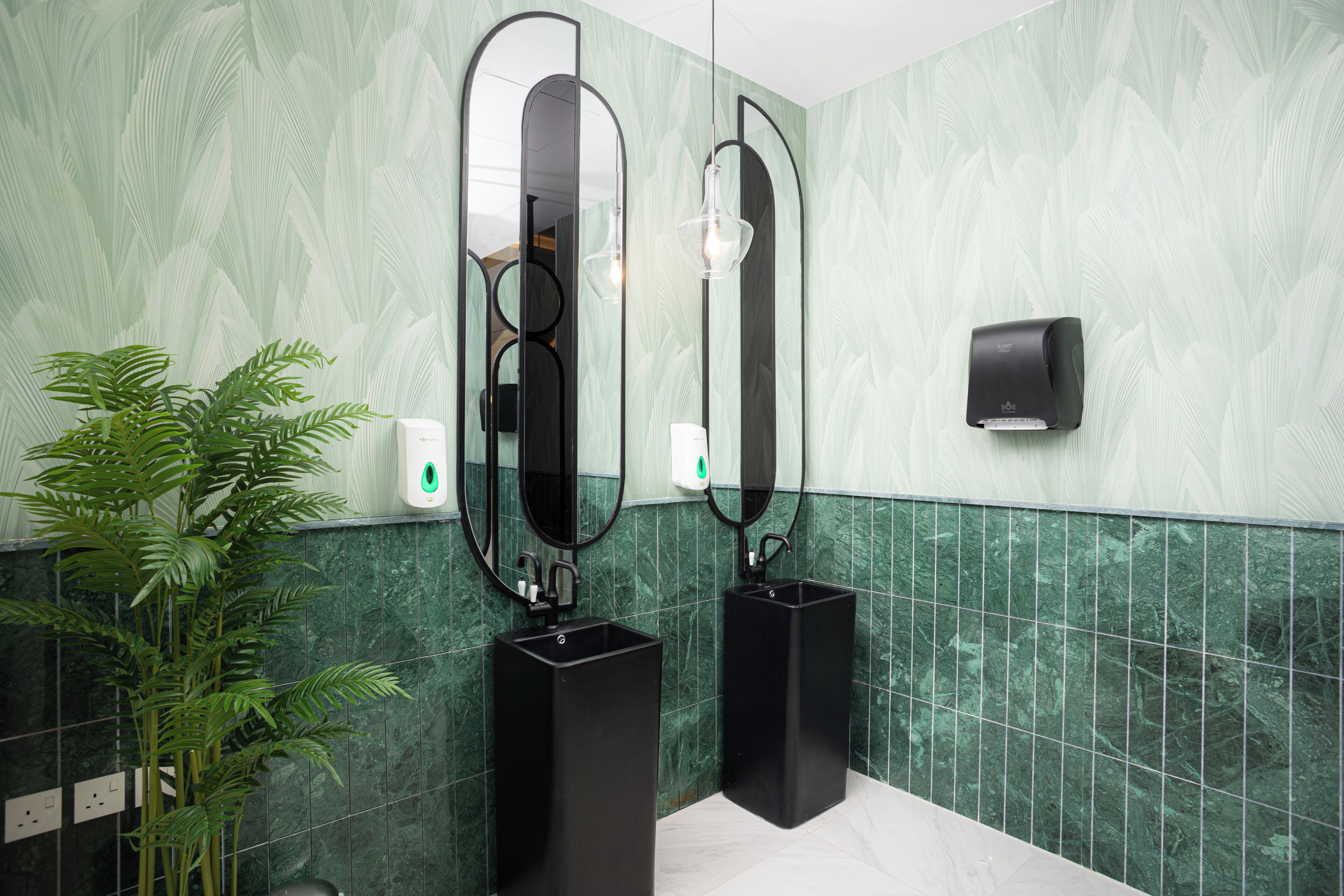 A bathroom with two sinks, a mirror, a hand dryer, and a potted plant.