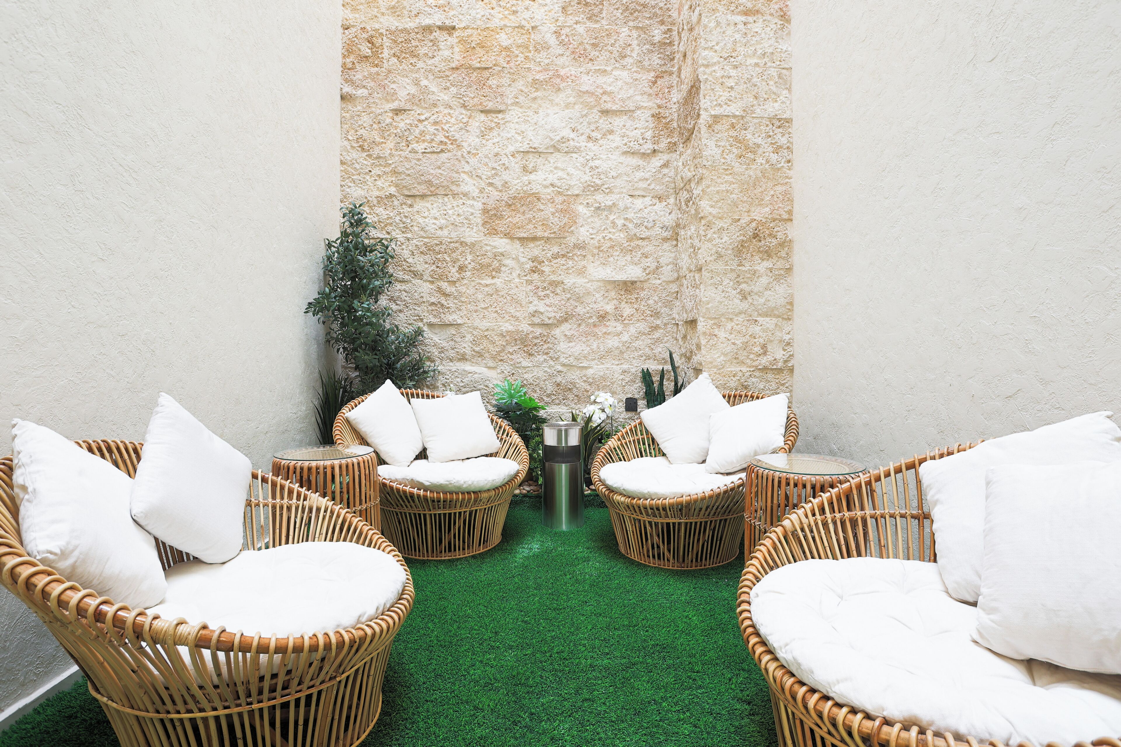 A corner with four wicker chairs with white cushions, a small round table with a metallic base, and a green artificial turf floor.