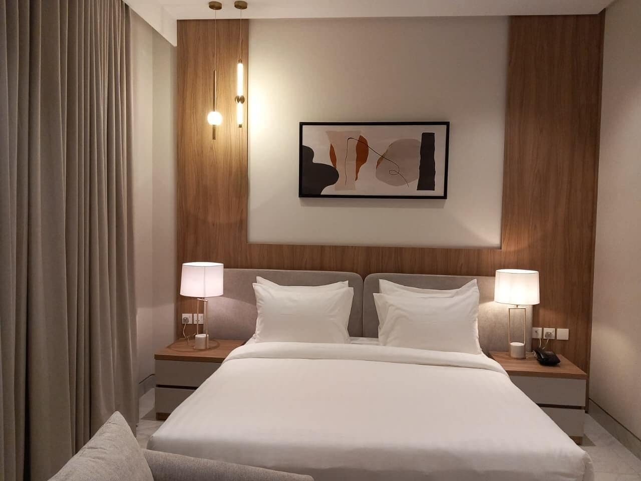 A modern hotel room with a large bed, two bedside tables with lamps, a wooden headboard, and a framed abstract artwork on the wall.