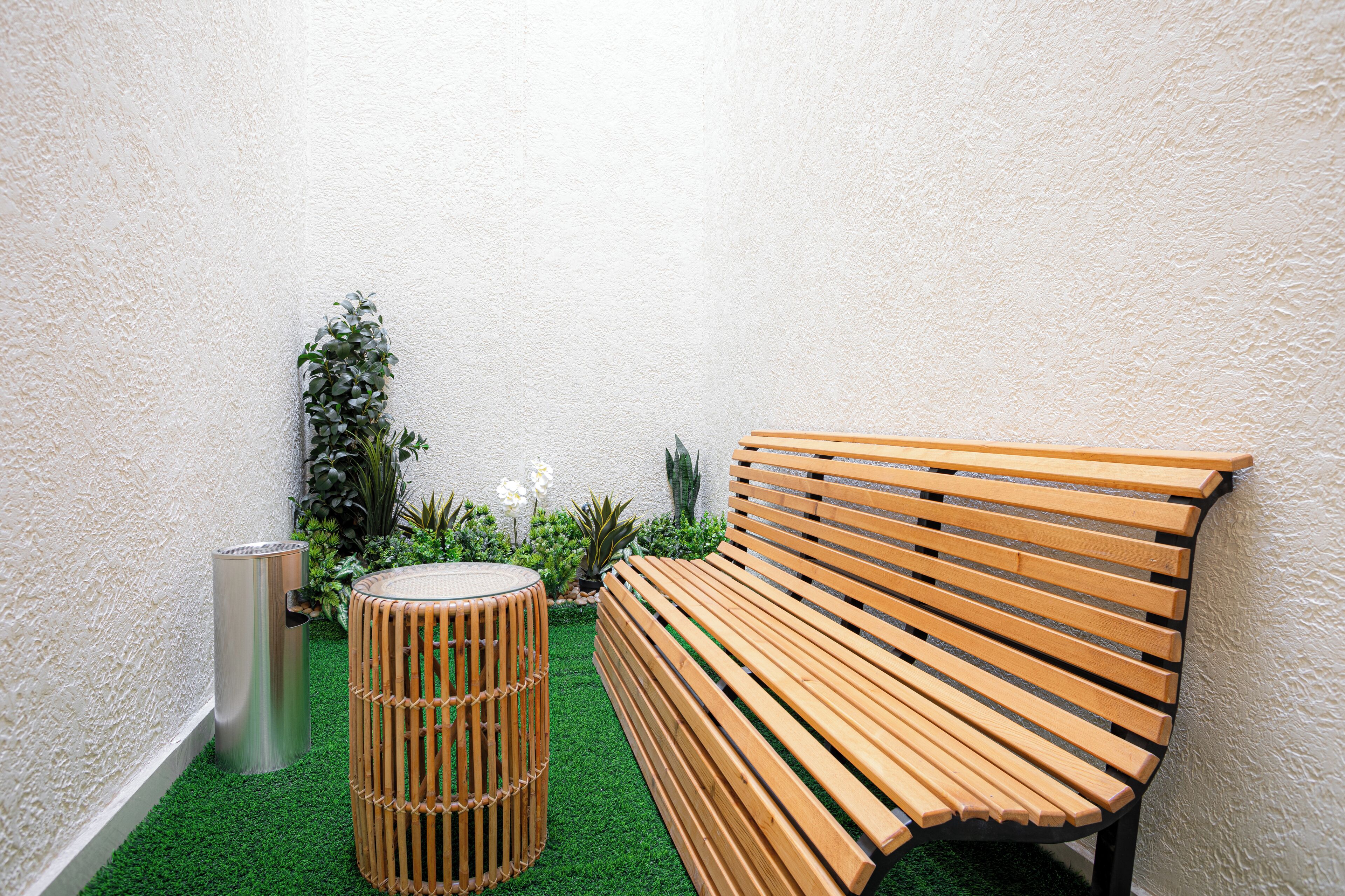 A wooden bench and a cylindrical stool in a small, enclosed space with artificial green turf and various plants.
