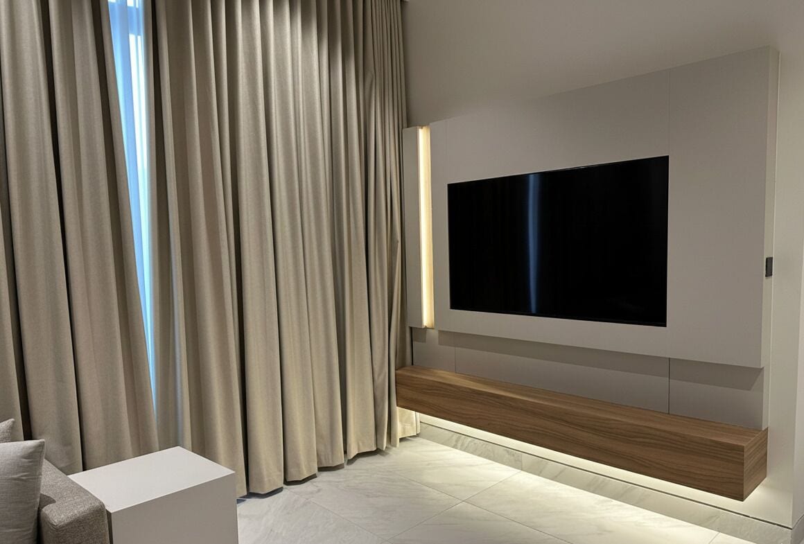 Un salon moderne avec un téléviseur à écran plat fixé au mur, un banc en bois et une petite table d’appoint blanche.
