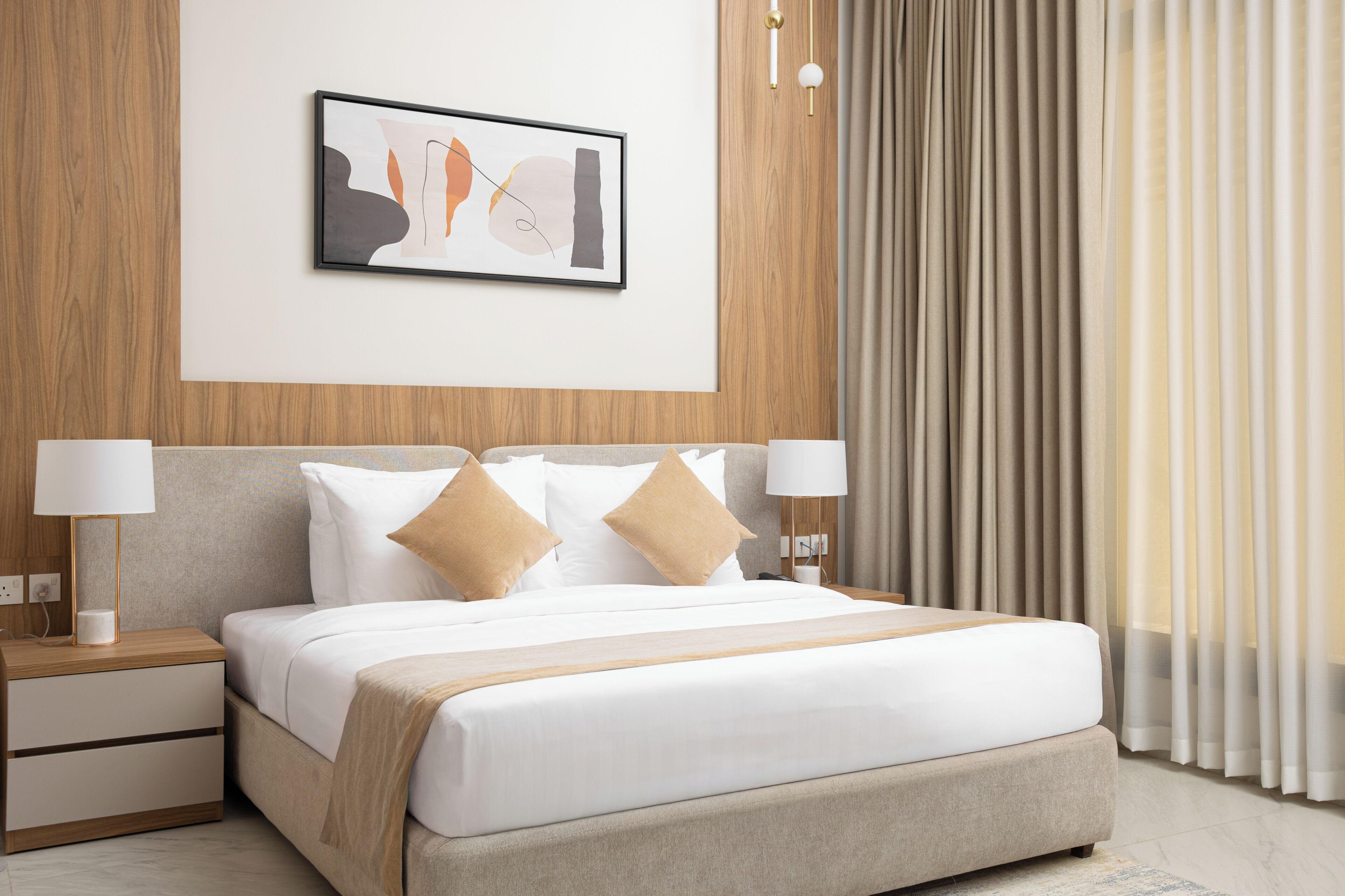 A modern hotel room with a bed, nightstands, a lamp, a framed abstract artwork, and a window with curtains.