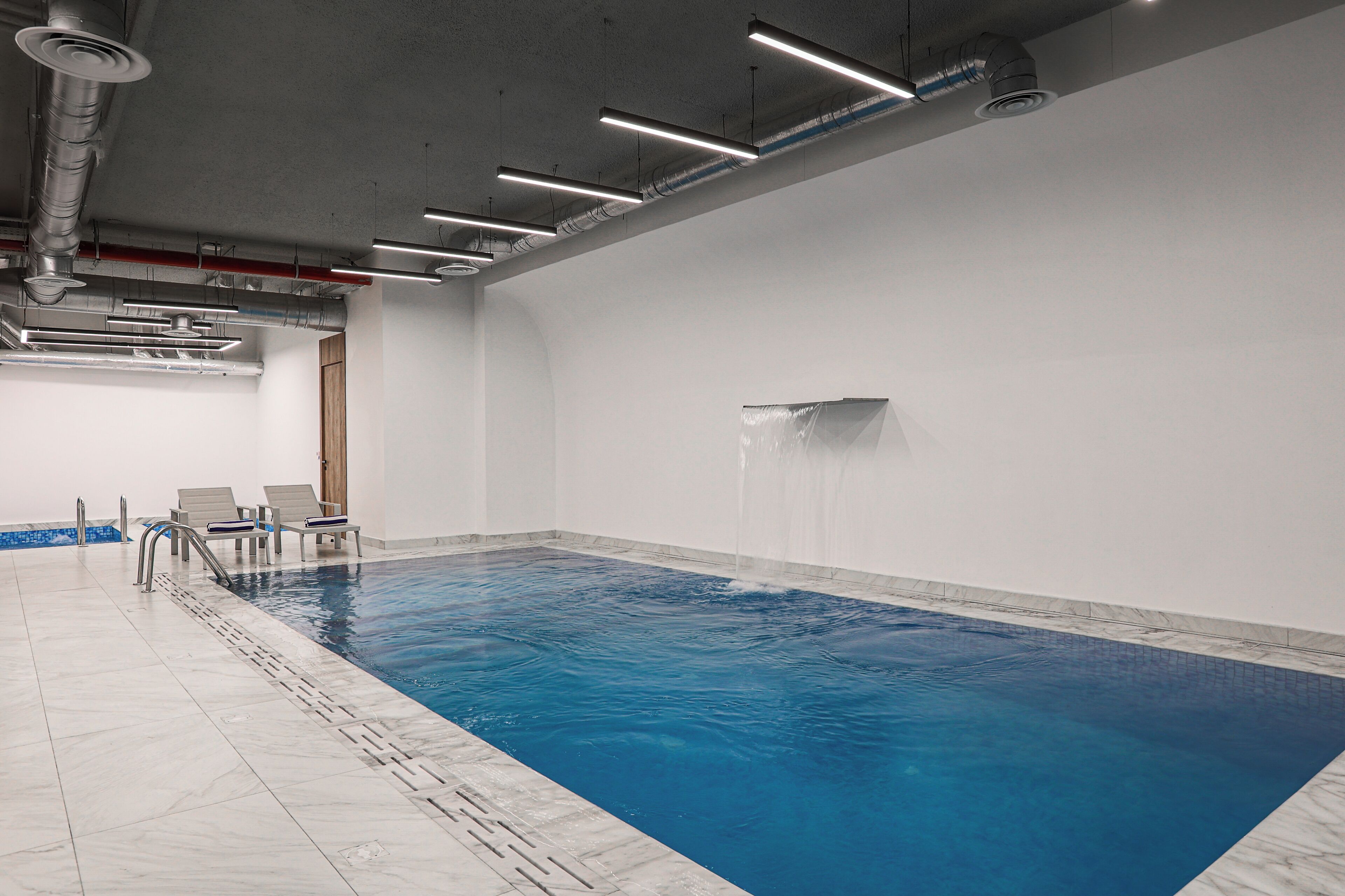 Ein Hallenbad mit klarem Wasser, gefliestem Boden und einer Decke mit freiliegenden Leitungen.