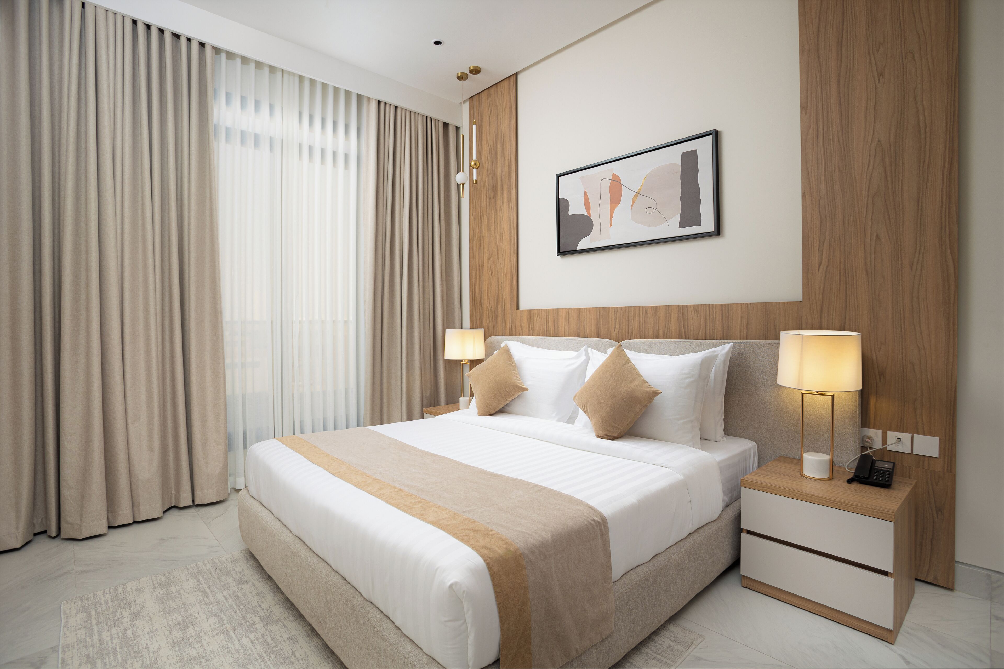 A modern hotel room with a large bed, two bedside tables, a nightstand, a framed abstract artwork, and sheer curtains.