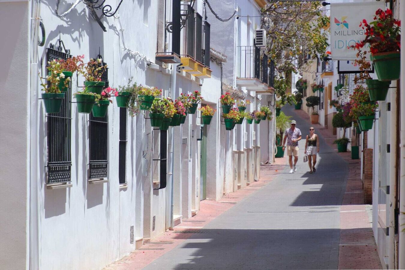 A narrow street lined with white buildings, each adorned with hanging flower baskets.