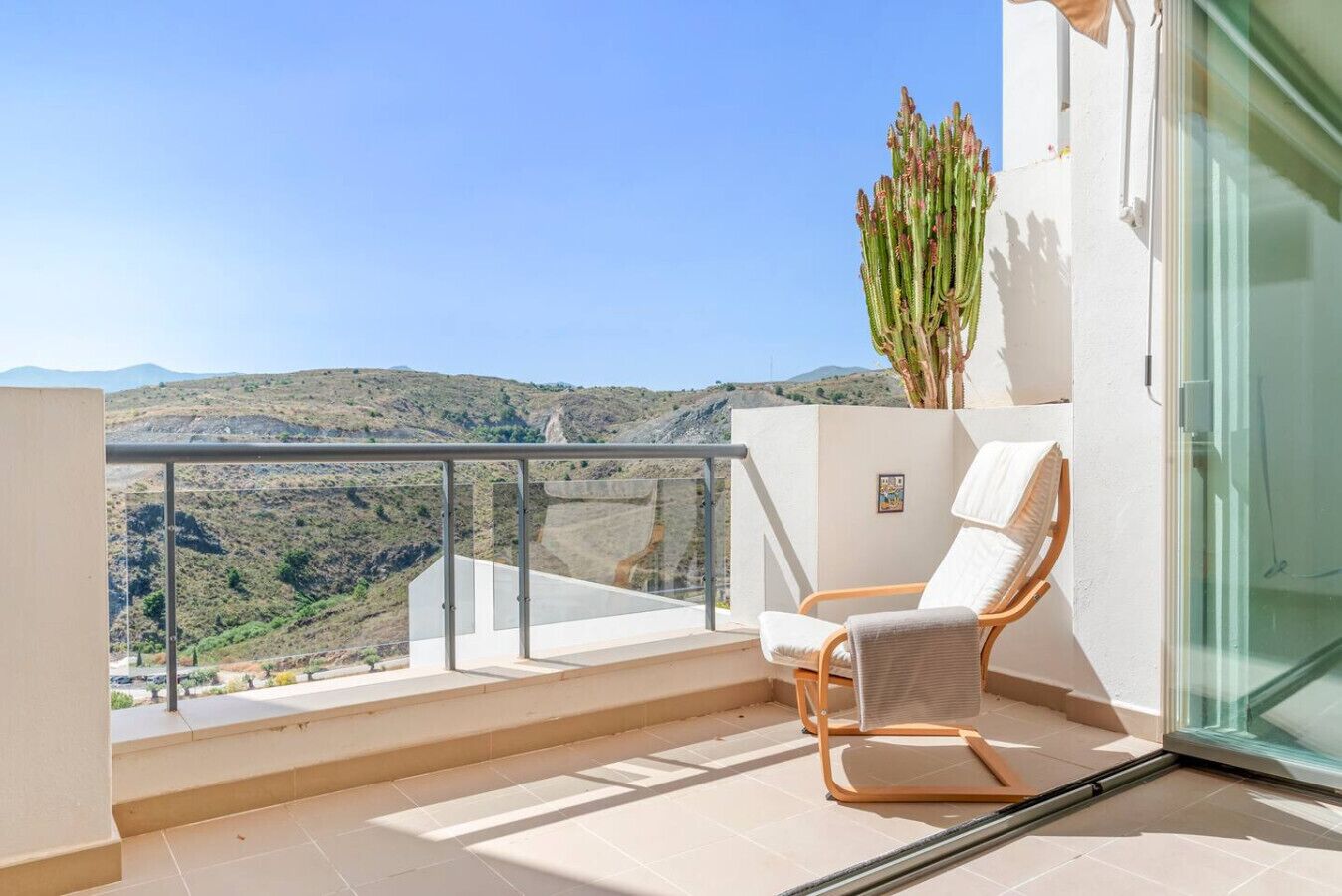 A balcony with a white chair, a cactus, and a view of a hilly landscape.