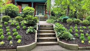 Un edificio residencial con puerta verde, un juego de escalones de hormigón que conducen a la entrada y un jardín bien cuidado con diferentes arbustos y plantas.