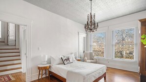 Un dormitorio con cama de madera, paredes blancas, una araña y ventanas grandes.