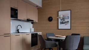 Una cocina moderna con armarios de madera, un grifo negro y un horno empotrado. Hay una mesa redonda de comedor con sillas grises y una alfombra negra. Un reloj y un cuadro enmarcado están en la pared.