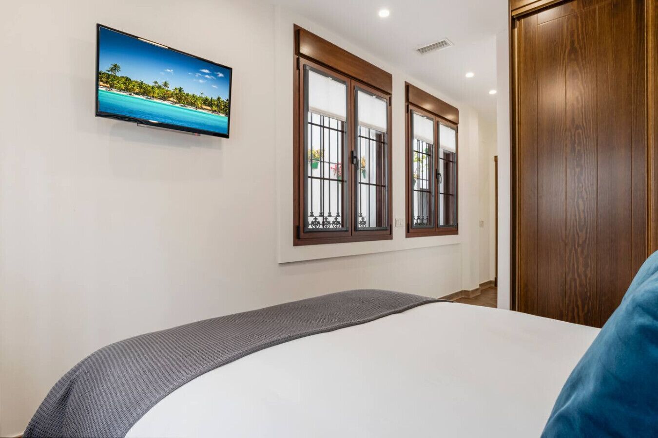 Un dormitorio con una cama, un televisor montado en la pared y dos ventanas con marcos de madera.