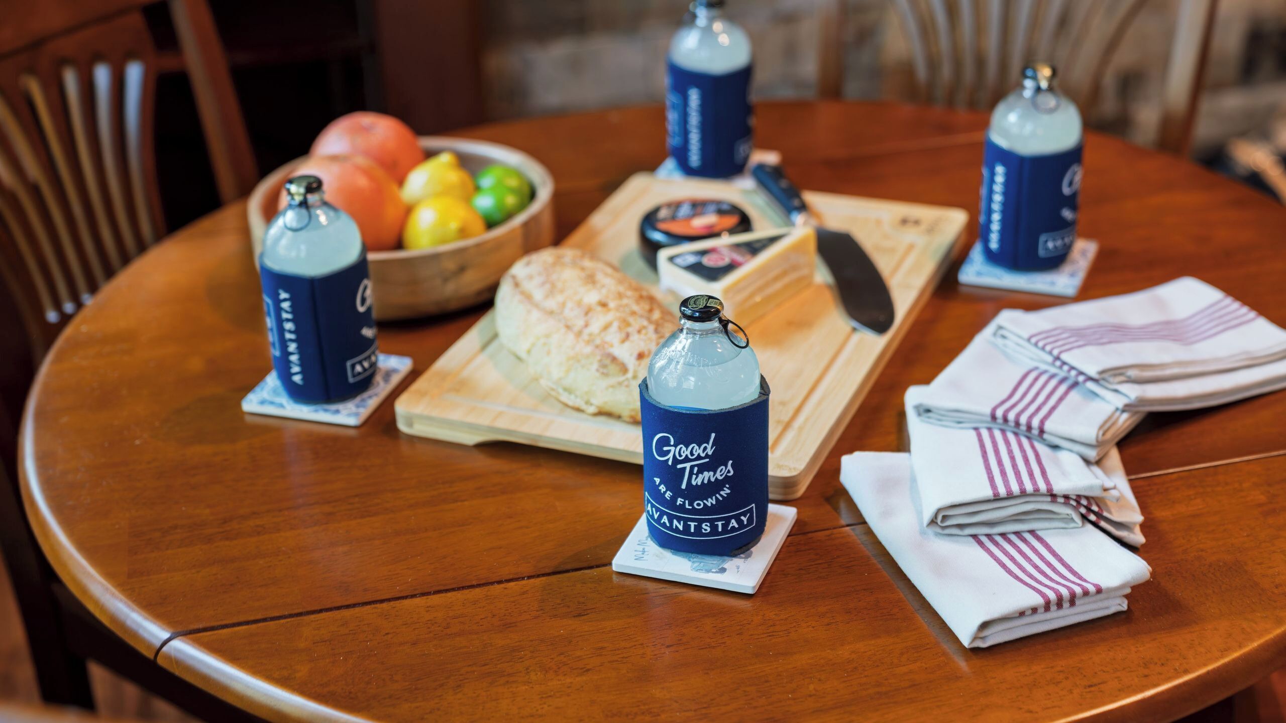 Una mesa redonda de madera con un cuenco de fruta, una hogaza de pan, una botella de agua y toallas dobladas.