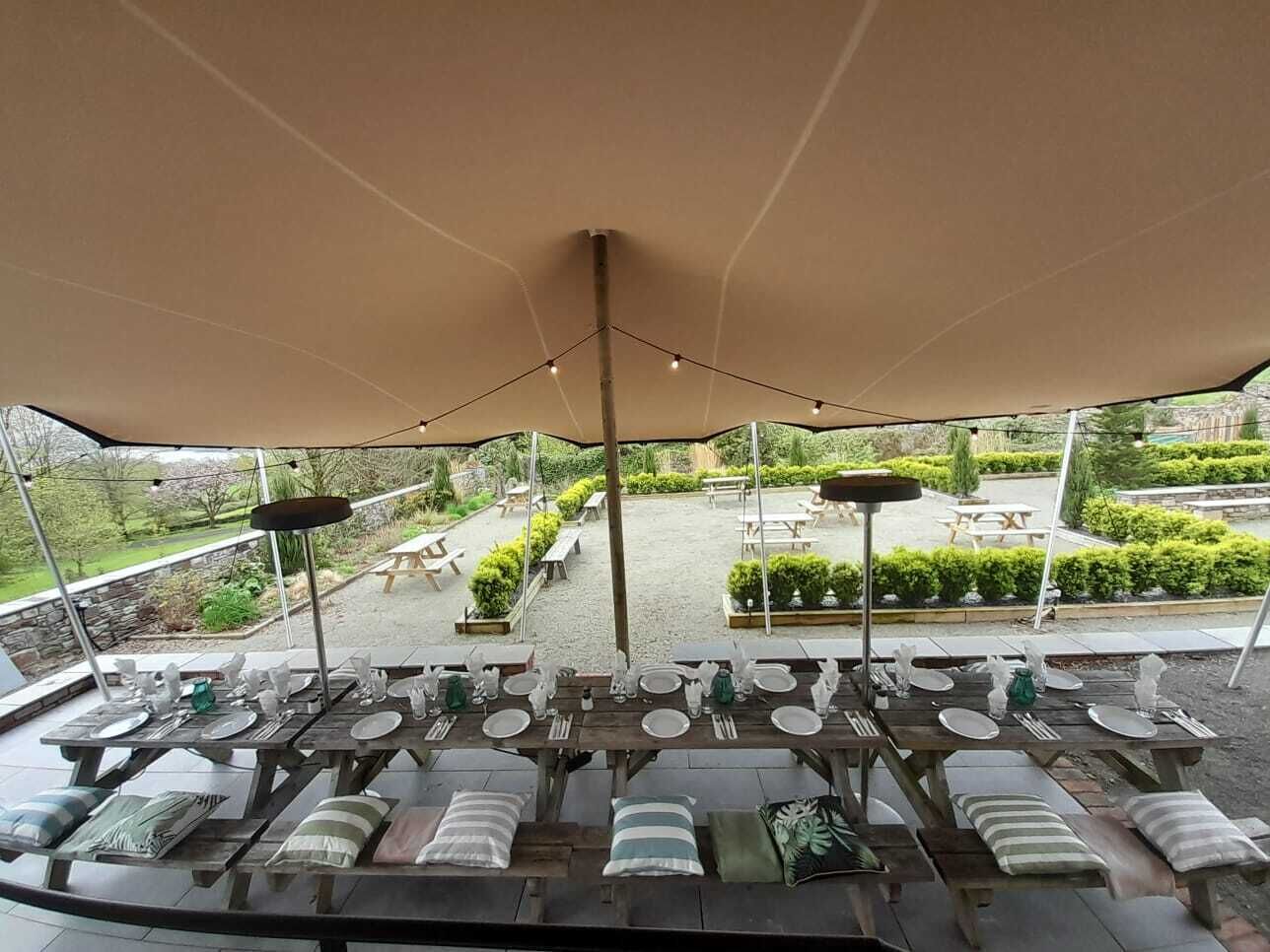 An outdoor dining area with tables and chairs under a large canopy, overlooking a garden with benches and picnic tables.
