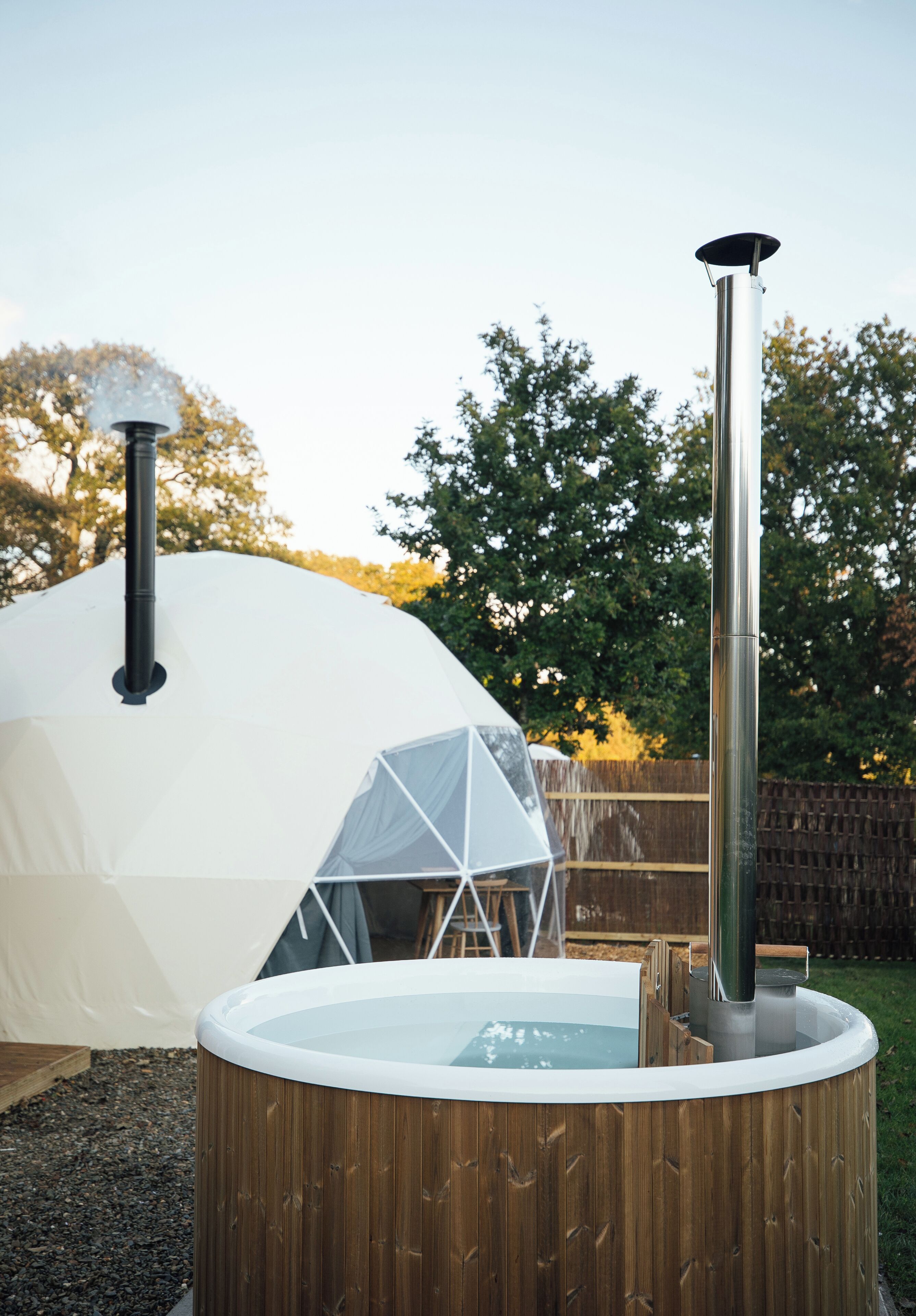 A hot tub with a wooden exterior and a chimney, a white tent-like structure, and a wooden fence in the background.
