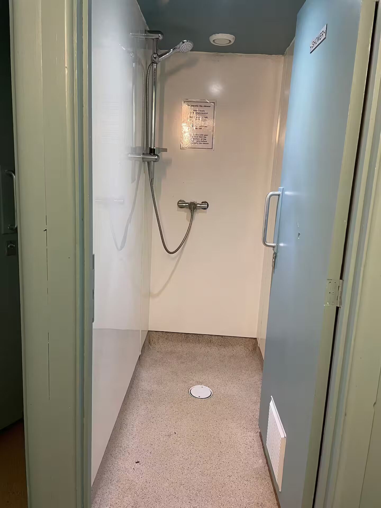 Une petite salle de douche étroite, équipée d’un pommeau de douche, d’un flexible de douche et d’un panneau de commande fixé au mur.