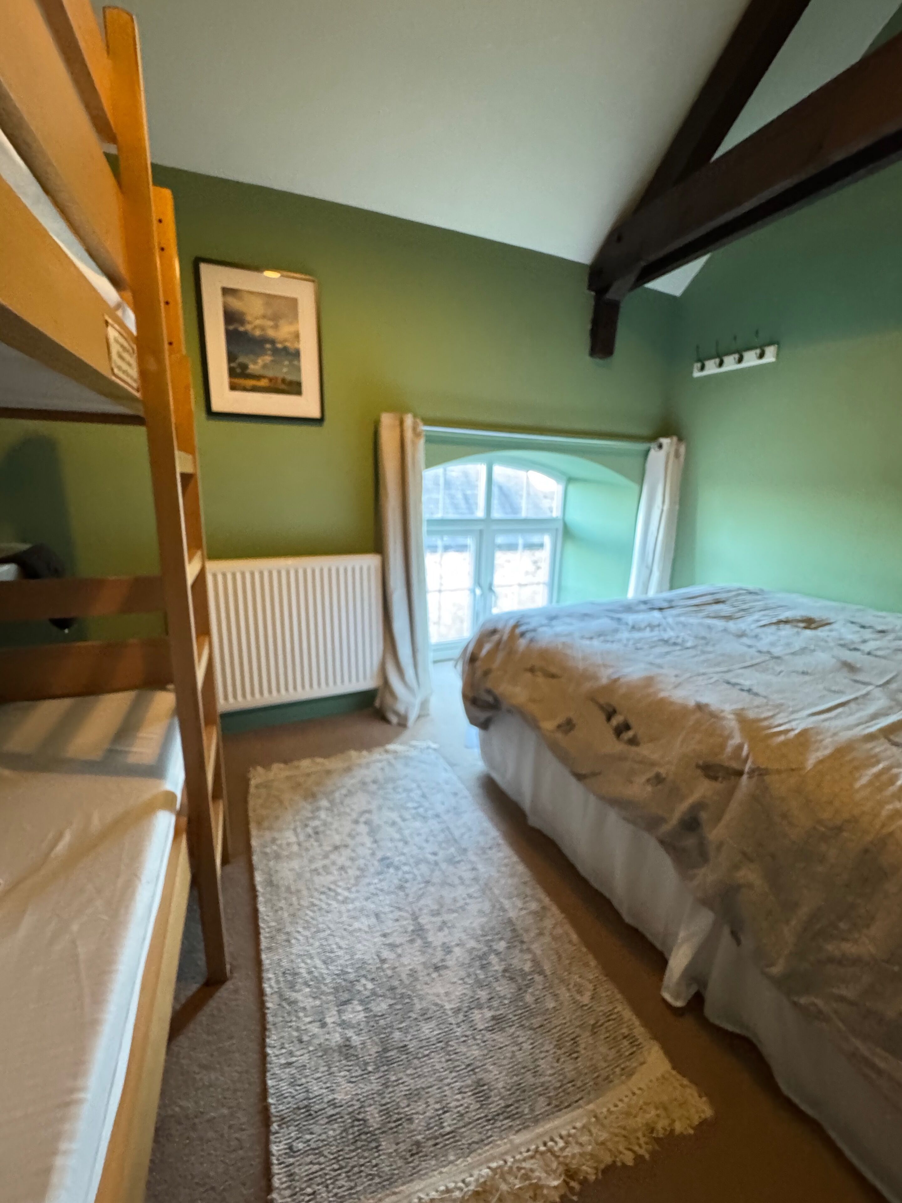 Una habitación con literas, una ventana con cortinas, un radiador y un cuadro en la pared.