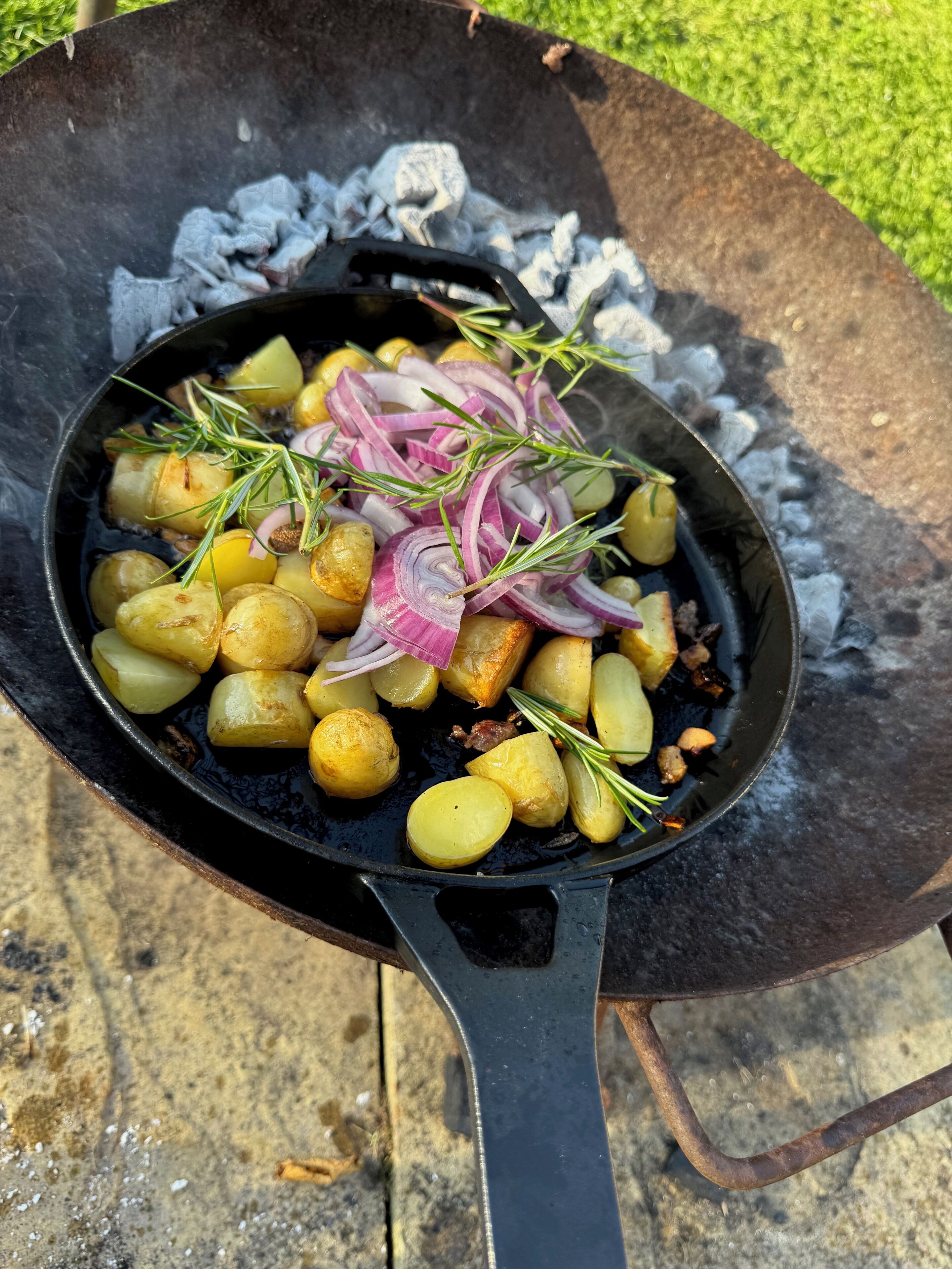 A cast iron skillet filled with roasted potatoes, red onions, and rosemary on a grill.