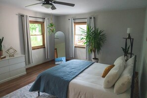A bedroom with a bed, a ceiling fan, a window, a mirror, a dresser, and a potted plant.