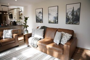 A living room with a brown leather sofa, a coffee table, and a patterned rug. There are three framed paintings on the wall depicting mountainous landscapes. - The Firefly Retreat in Lancaster county (Manheim)