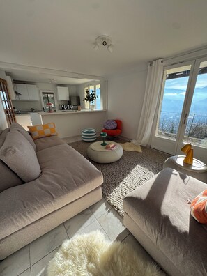 Uma sala de estar moderna com um sofá, um banquinho e vista para as montanhas por uma grande janela.
