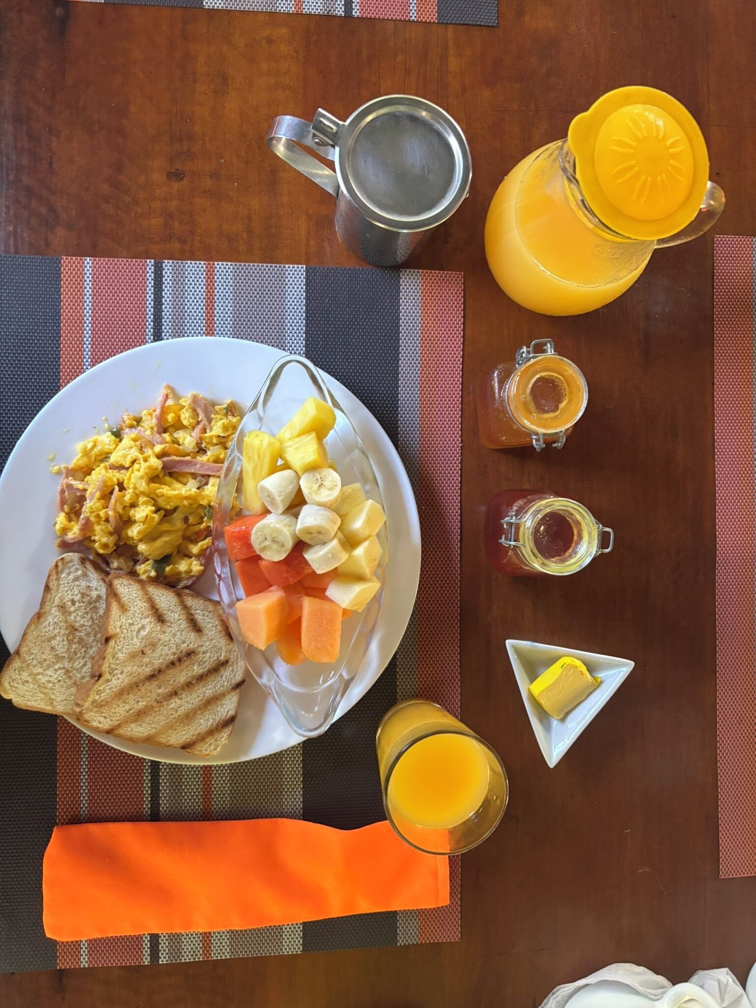 Una mesa de desayuno con huevos revueltos, fruta, tostadas, una cafetera, una jarra de jugo y un vaso de zumo de naranja.