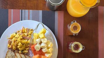 Una mesa de desayuno con huevos revueltos, fruta, tostadas, una cafetera, una jarra de jugo y un vaso de zumo de naranja.