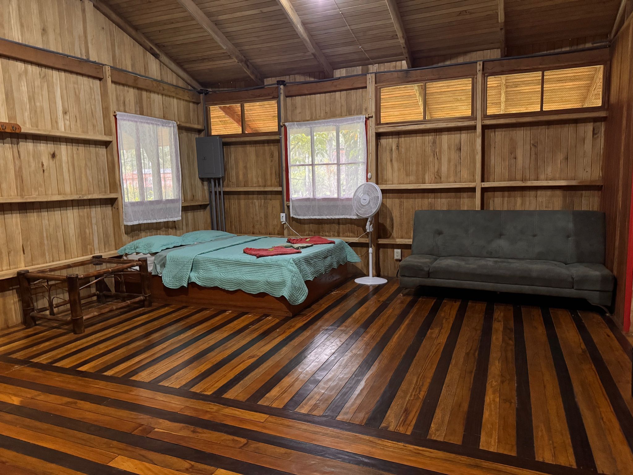 A room with wooden walls and floors, a bed with a green bedspread, a grey sofa, and a standing fan.