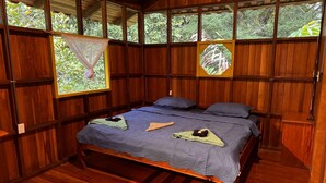 A wooden room with a bed, two pillows, and a window with a view of greenery. - Sabalo lodge (Sierpe)