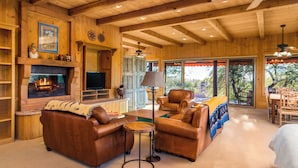 A cozy wooden cabin interior with a fireplace, leather sofas, a dining area, and large windows.