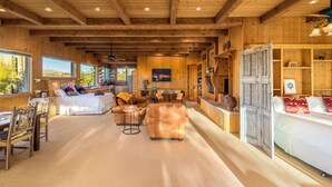 A spacious living area with a large sofa, a coffee table, and a dining table with chairs. There is a wooden ceiling with exposed beams and a large window providing a view of the outdoors.