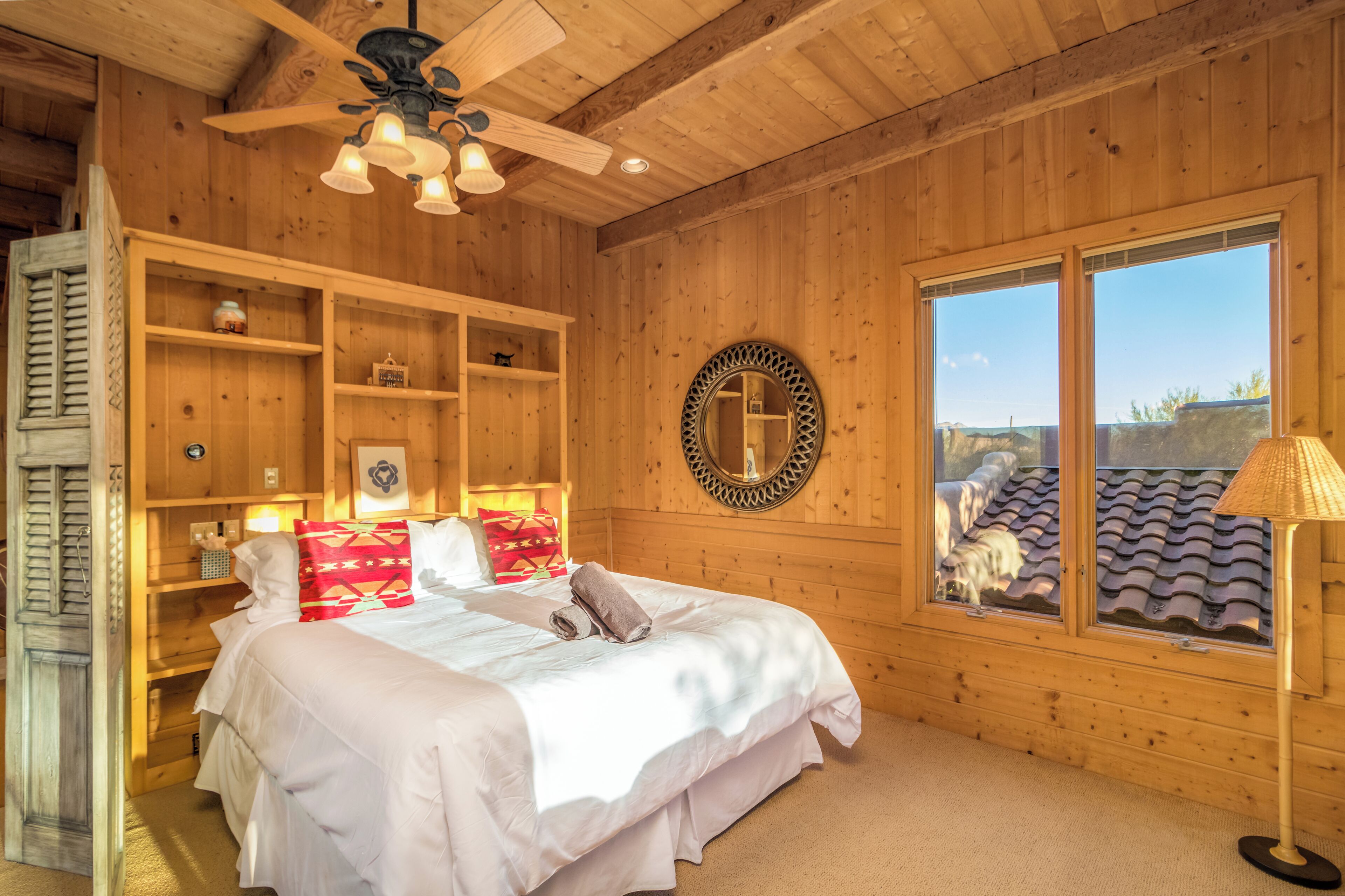 A wooden room with a bed, shelves, a mirror, and a window with a view of a dog and a tiled roof.