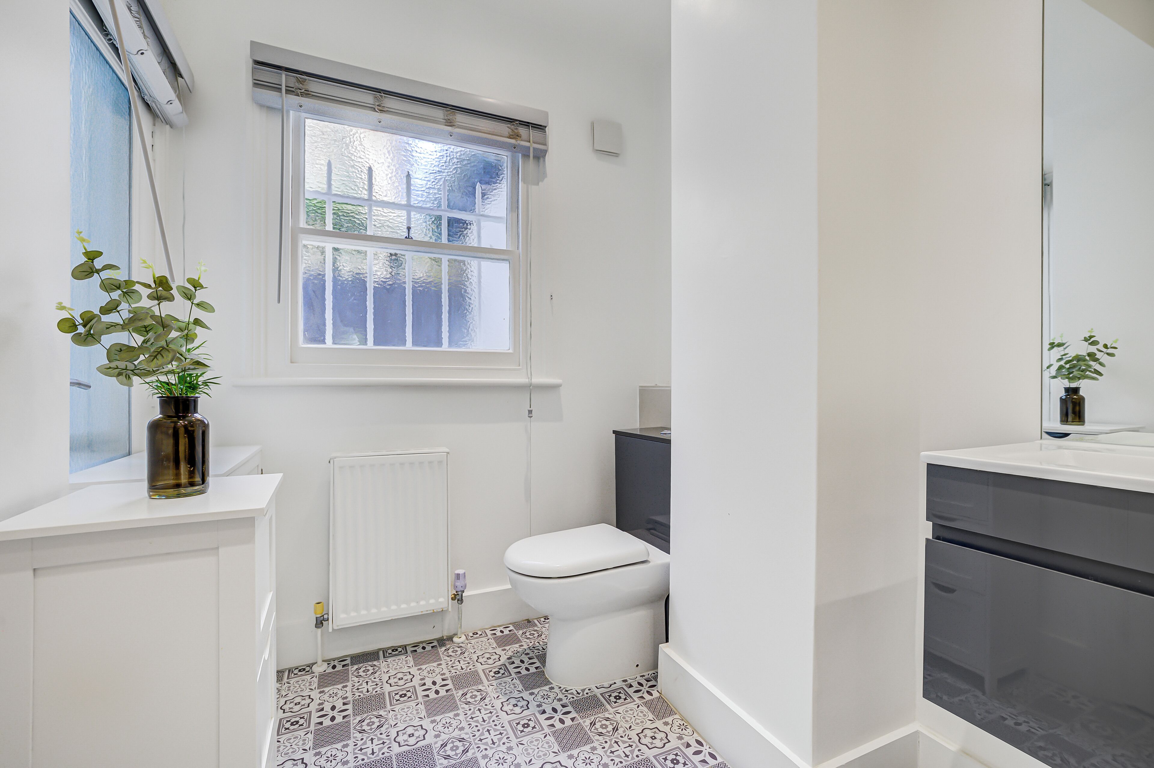 Une salle de bain avec des toilettes blanches, une fenêtre donnant sur un paysage agréable, un radiateur et un carrelage décoratif.