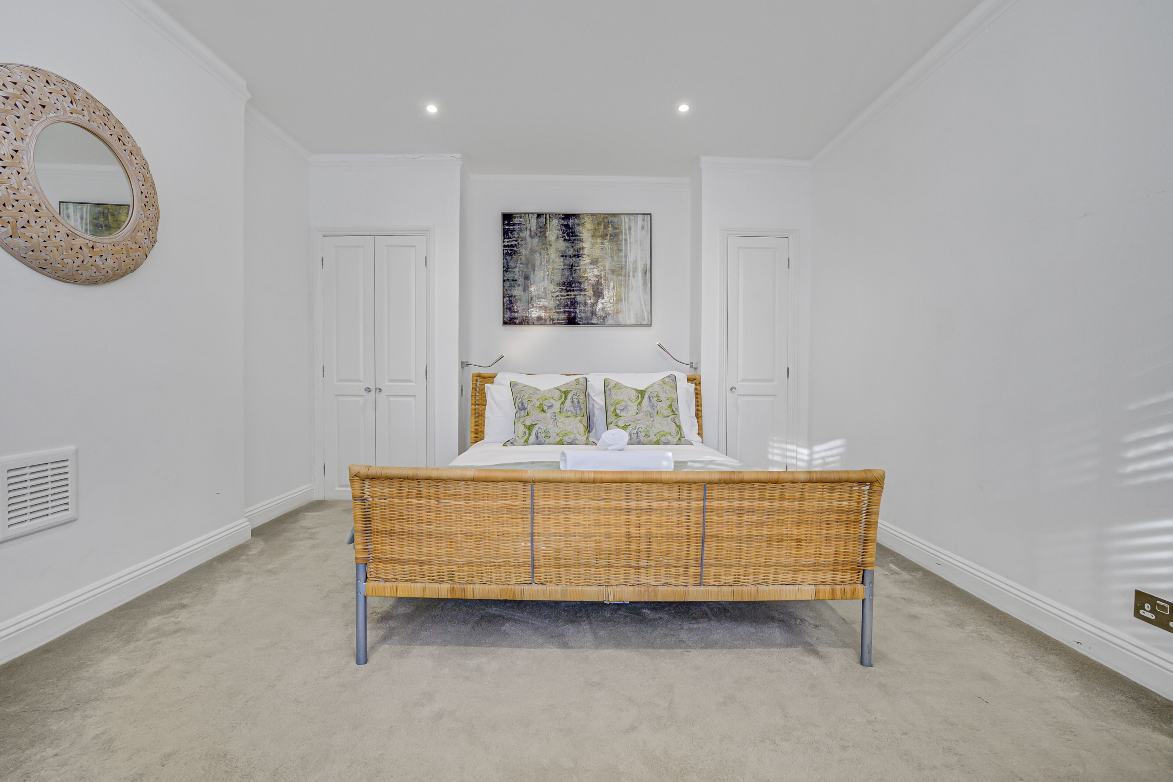 A bedroom with a wicker bed, white walls, a round mirror, and a painting.