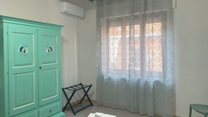 A room with teal-colored wardrobe, a black drying rack, a window with patterned curtains, and a white air conditioning unit. - Just minutes from Ogliastra's most beautiful beaches, rooms with en suite facilities (Cardedu)