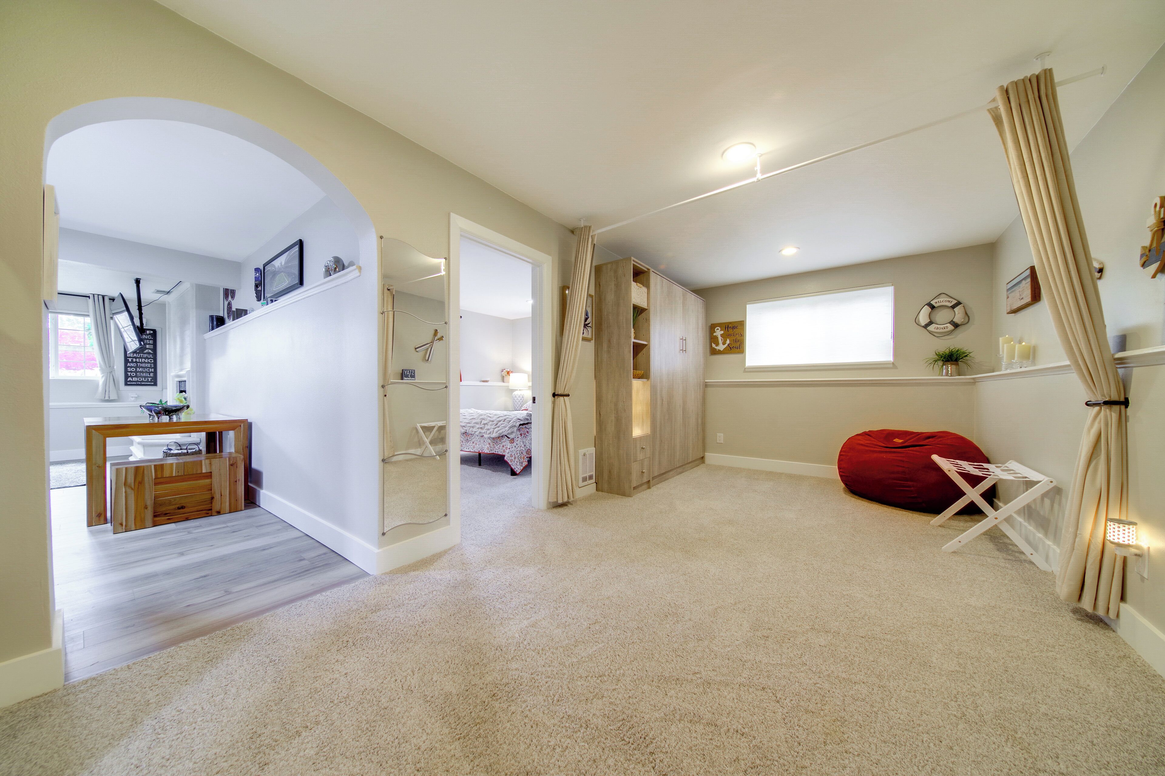 A spacious room with a large bed, a red bean bag, and a small table. There is a wooden desk with a chair and a mirror on the wall.