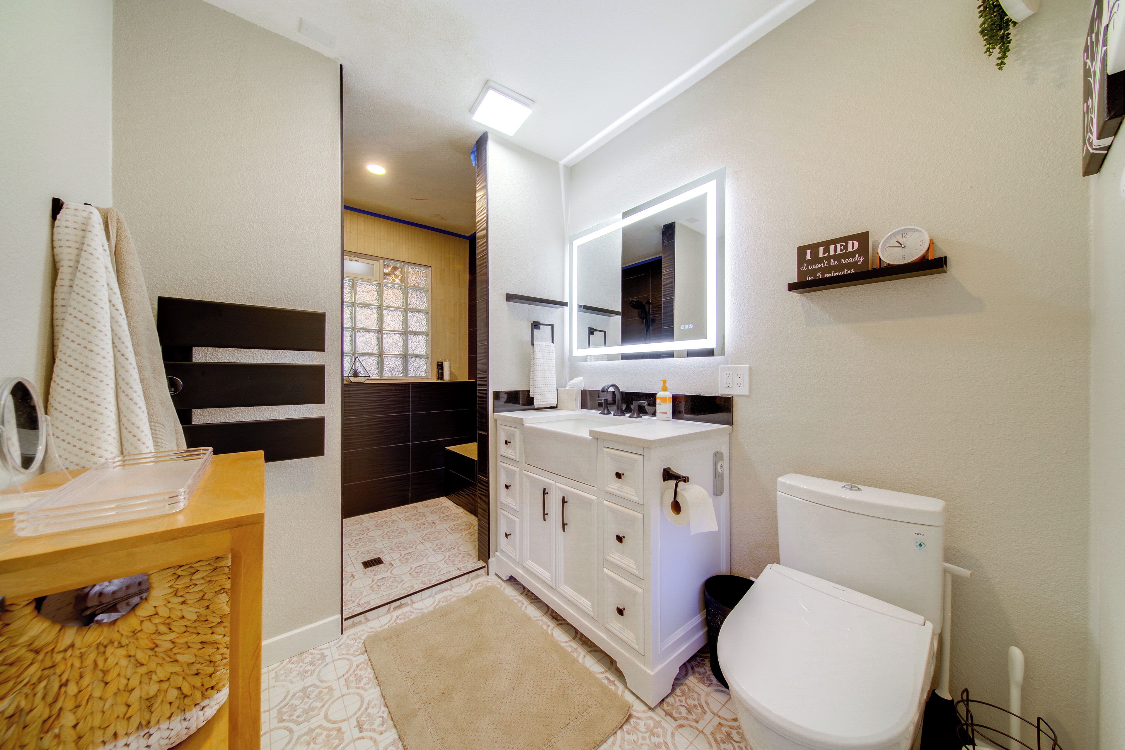 A modern bathroom with a white sink, mirror, and toilet. There is a wooden vanity with a towel rack and a clear glass tray. A patterned rug is on the floor.