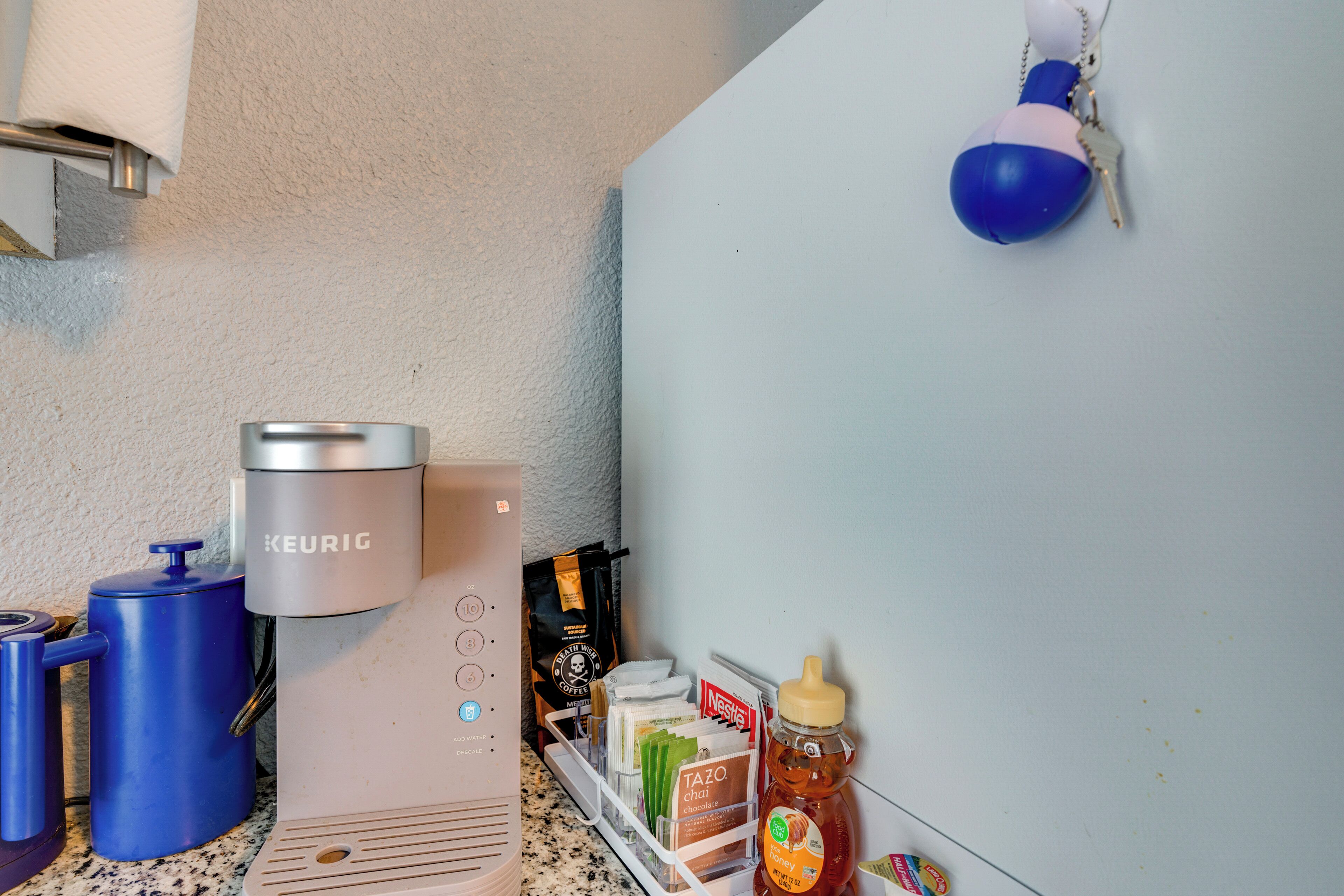 Pieni keittiönurkkaus, jossa on Keurig-kahvinkeitin, sininen vedenannostelija, kahvimylly, kahvikapselihylly, hunajapurkki ja teepussiannostelija.