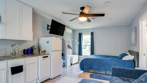 A modern hotel room with a kitchenette, a bed with blue bedding, a ceiling fan, and a large window with curtains. - 10 Mi to Lake Conroe: Couple’s Retreat w/ Porch! (Montgomery)