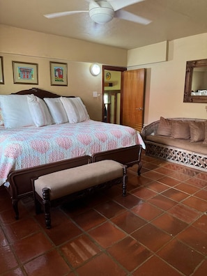 Ein Schlafzimmer mit einem Bett, einer Sitzbank, einer hölzernen Sitzbank, einem Deckenventilator, einem Spiegel und einer Tür.