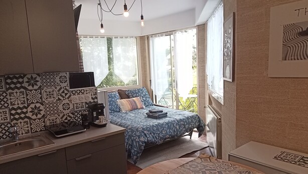 Habitació moderna i compacte, amb zona de cuina, llit amb roba de llit estampada i grans finestres que donen vistes a vegetació.