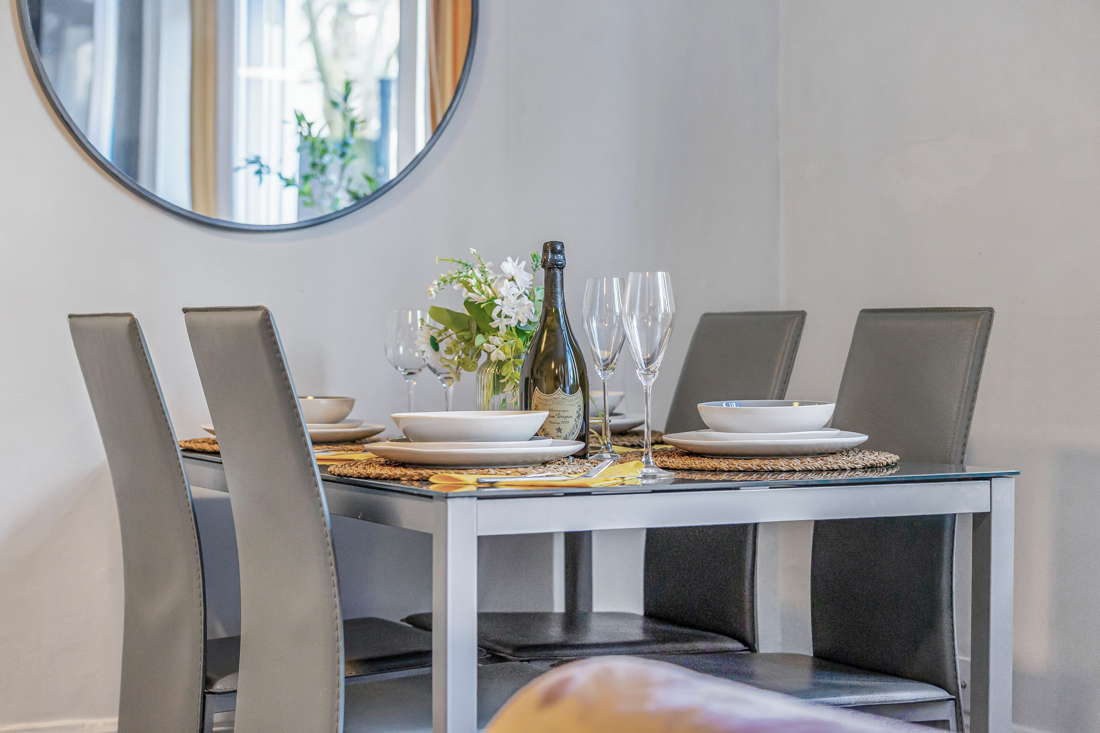 Une table à manger dressée avec de l’argenterie blanche, une bouteille de champagne et des verres, entour ée de chaises à revêtement gris.
