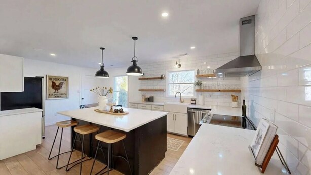 Una cocina moderna con armarios blancos, una isla central con taburetes, iluminación colgante y una campana extractora de acero inoxidable.