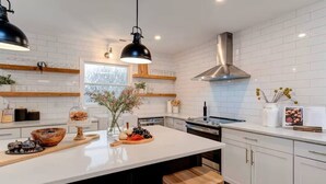 Una cocina moderna con azulejos blancos tipo metro, estantes de madera, una isla central con taburetes e iluminación colgante.