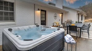 Un jacuzzi en un espacio exterior cubierto, con un muro de ladrillo, una mesa con sillas y un rincón de descanso con cojines.