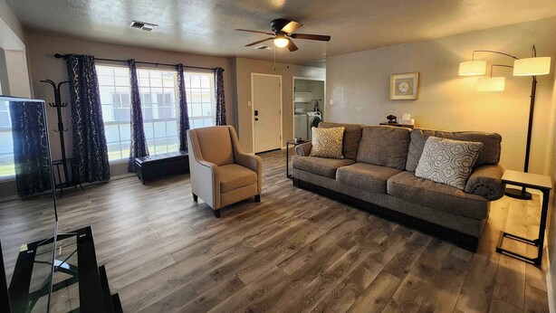A living room with a sofa, armchair, and a ceiling fan. There is a window with curtains, a door, and a lamp. - Modern Stay Near Fort Sill + Park & Shopping (Lawton)