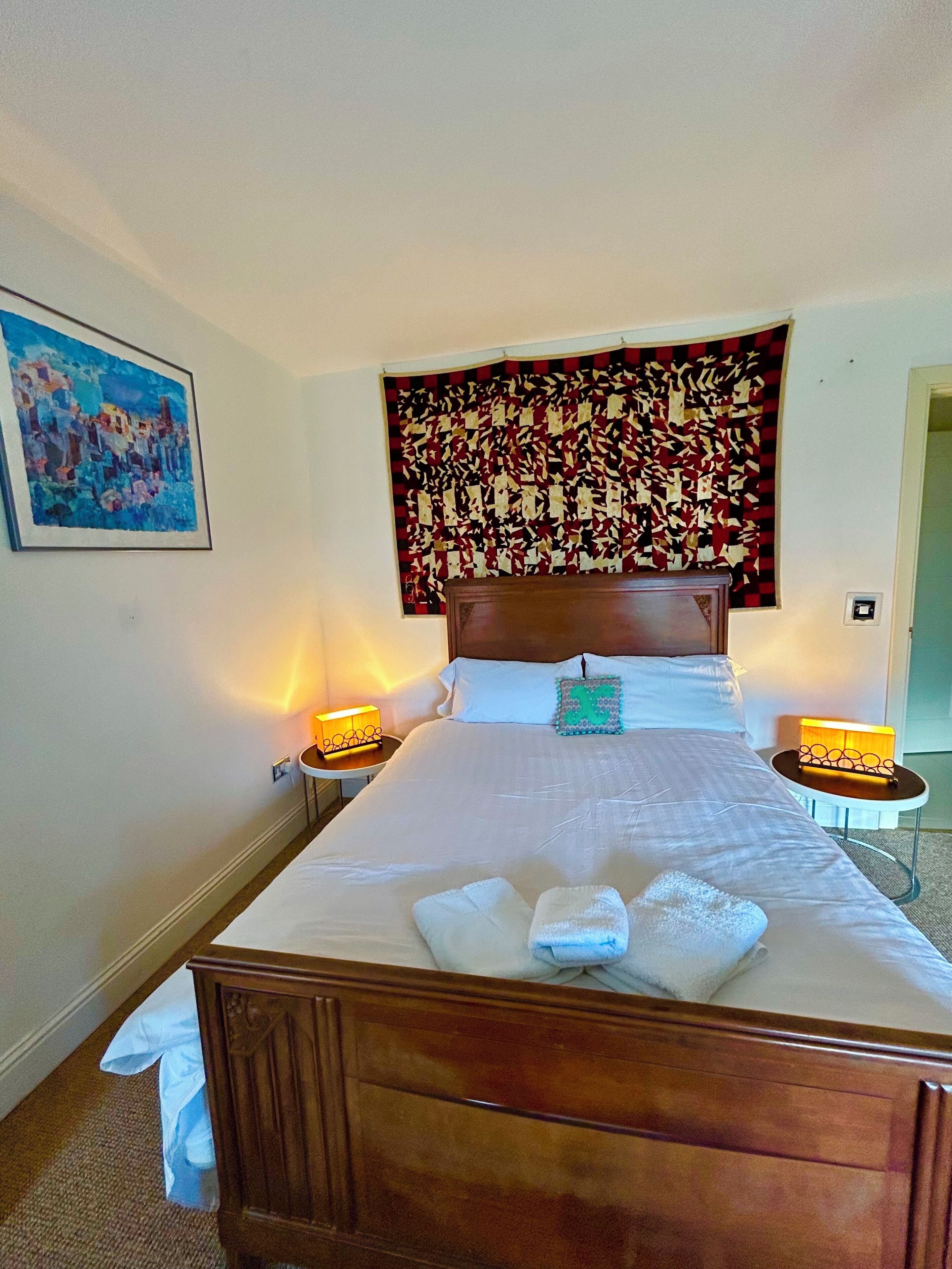 A neatly made bed with white linens and towels, two bedside lamps, a headboard with a patterned tapestry, and a painting on the wall.