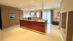 Una cocina moderna con una isla central, horno empotrado, luces colgantes y un amplio ventanal con cortinas.